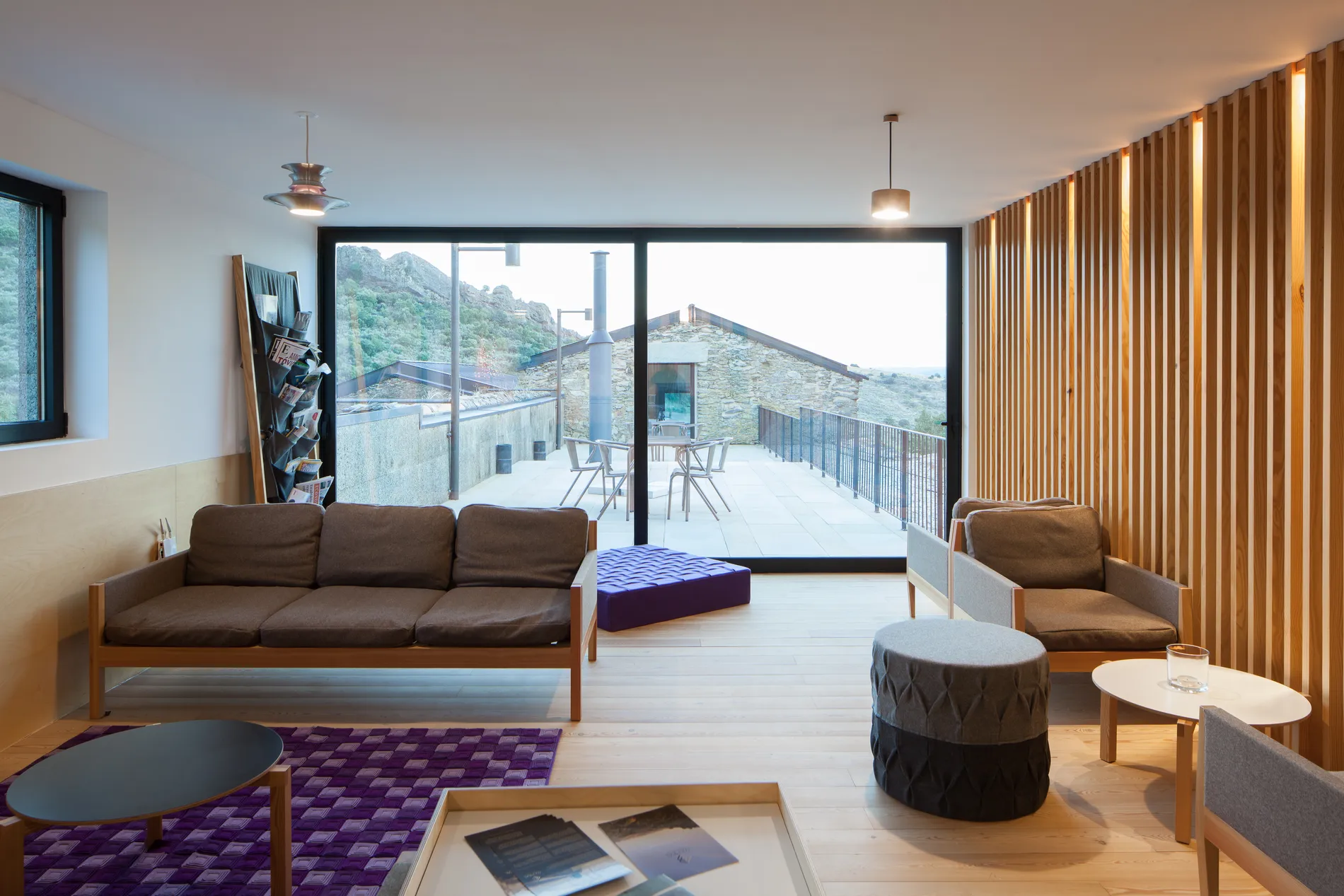 Sala moderna com sof&aacute;, poltrona, mesas e vista para &aacute;rea externa com montanhas.