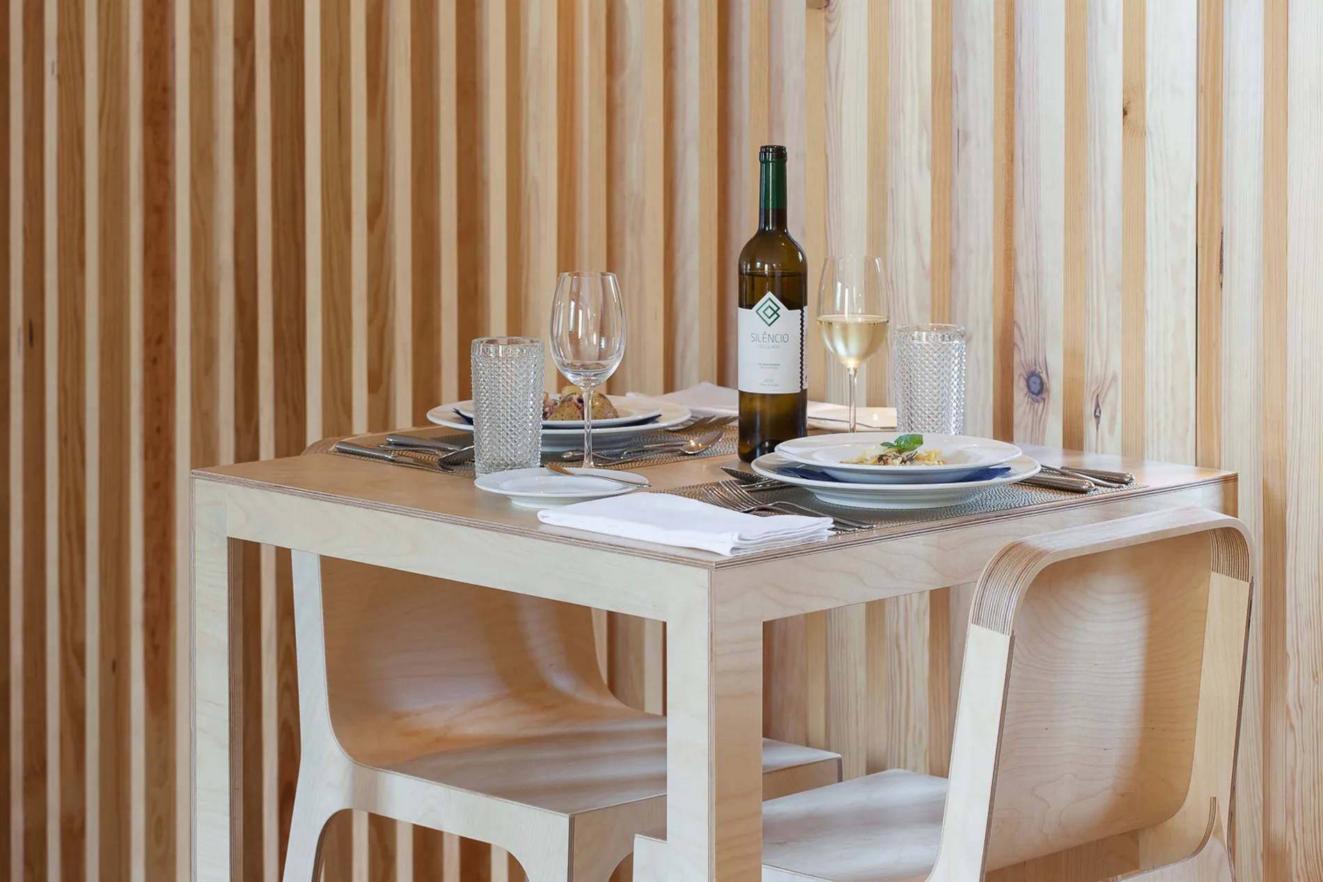 Mesa de madeira com pratos, talheres, ta&ccedil;as de vinho e garrafa, cercada por parede de ripas.