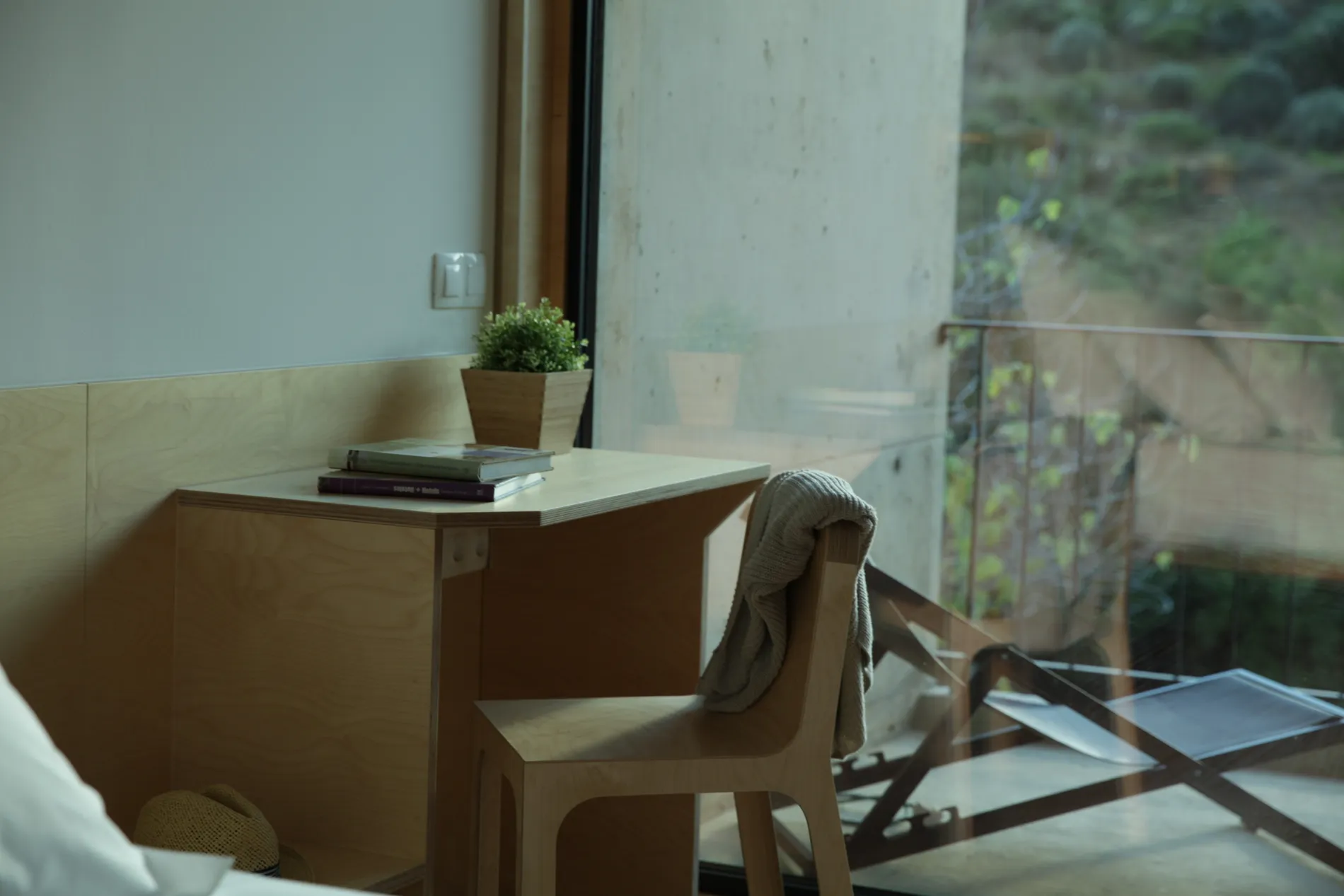 Quarto minimalista com mesa, cadeira, su&eacute;ter, plantas e vista para um jardim pela janela.