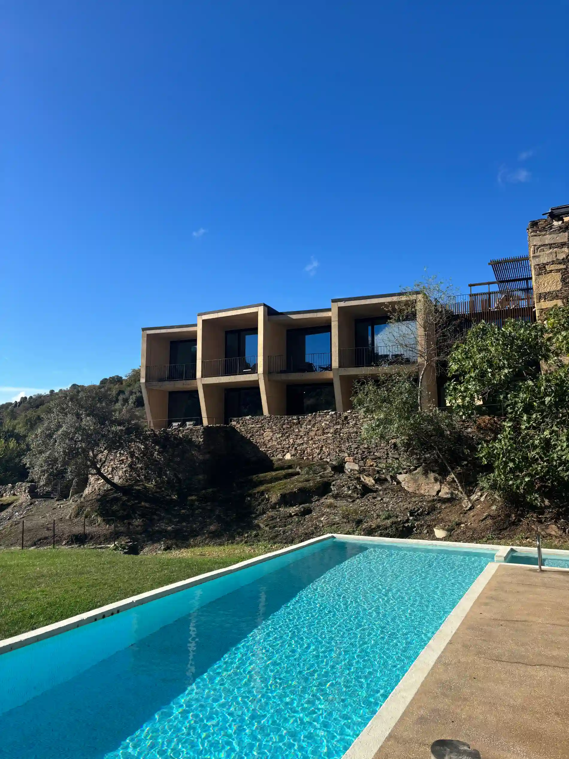 Casa moderna em colina, com piscina azul em primeiro plano e c&eacute;u claro ao fundo.