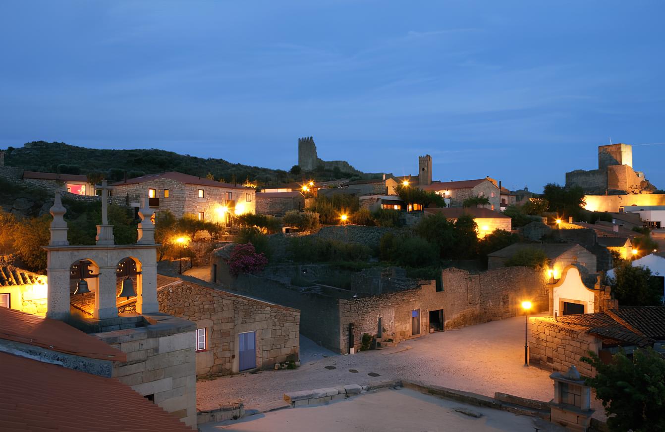 Vista noturna de uma vila iluminada com edif&iacute;cios e torres antigas ao fundo.