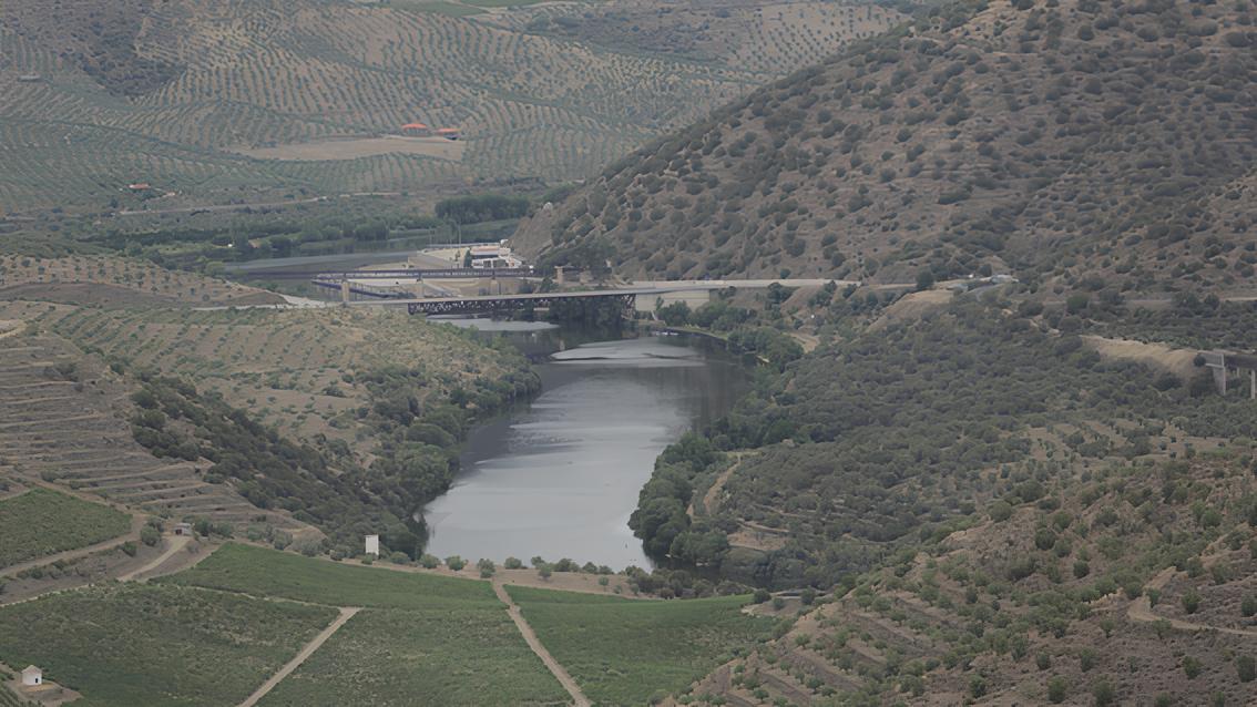 Paisagem montanhosa com rio ao centro e ponte ao fundo, cercada por vegeta&ccedil;&atilde;o e terra&ccedil;os.