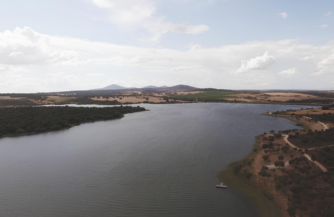 Lago e paisagem rural com &aacute;rvores, campos e montanhas ao fundo sob c&eacute;u com nuvens.