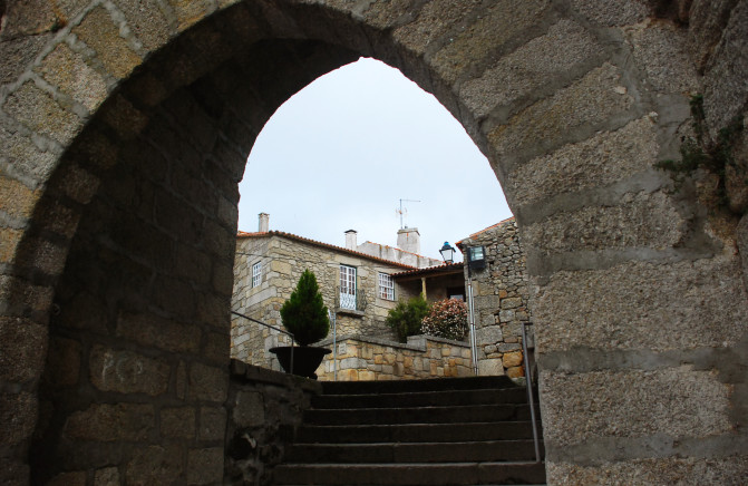 Arco de pedra com escadas levando a casas de pedra e vasos de plantas ao fundo.