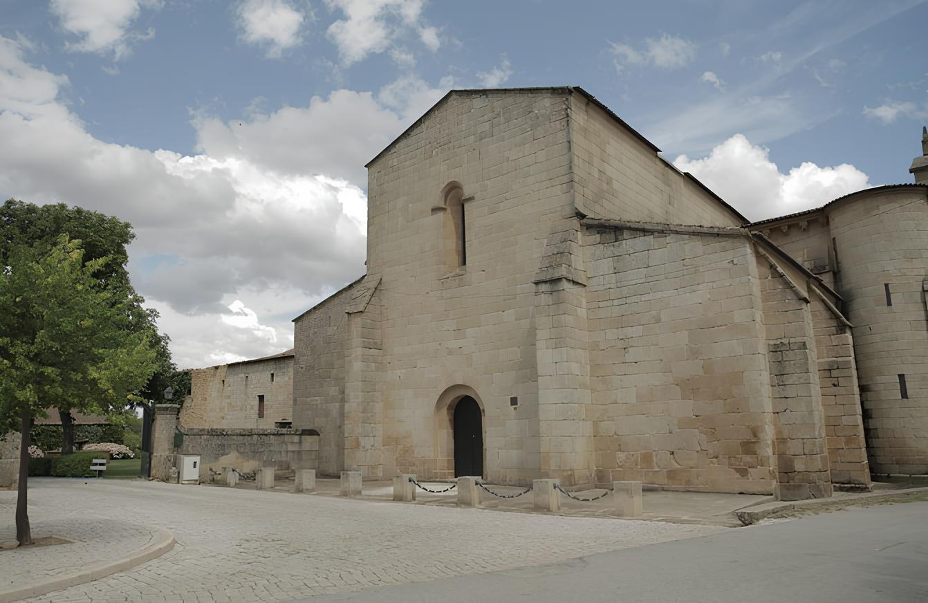 Igreja antiga de pedra sob um c&eacute;u parcialmente nublado, com &aacute;rvore e rua pavimentada ao lado.
