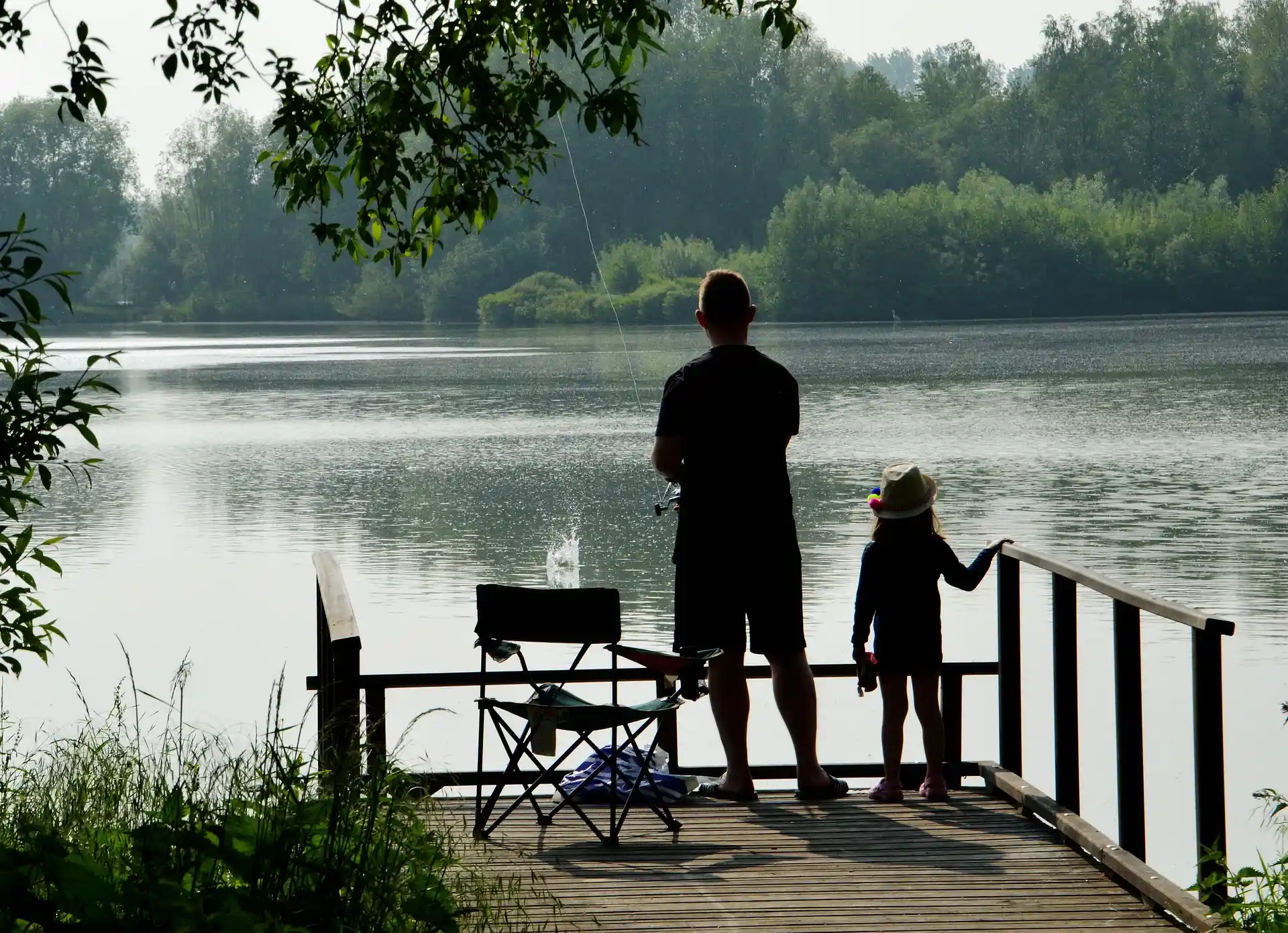 Pessoa e crian&ccedil;a pescando em um lago, com cadeiras e &aacute;rvores ao redor.