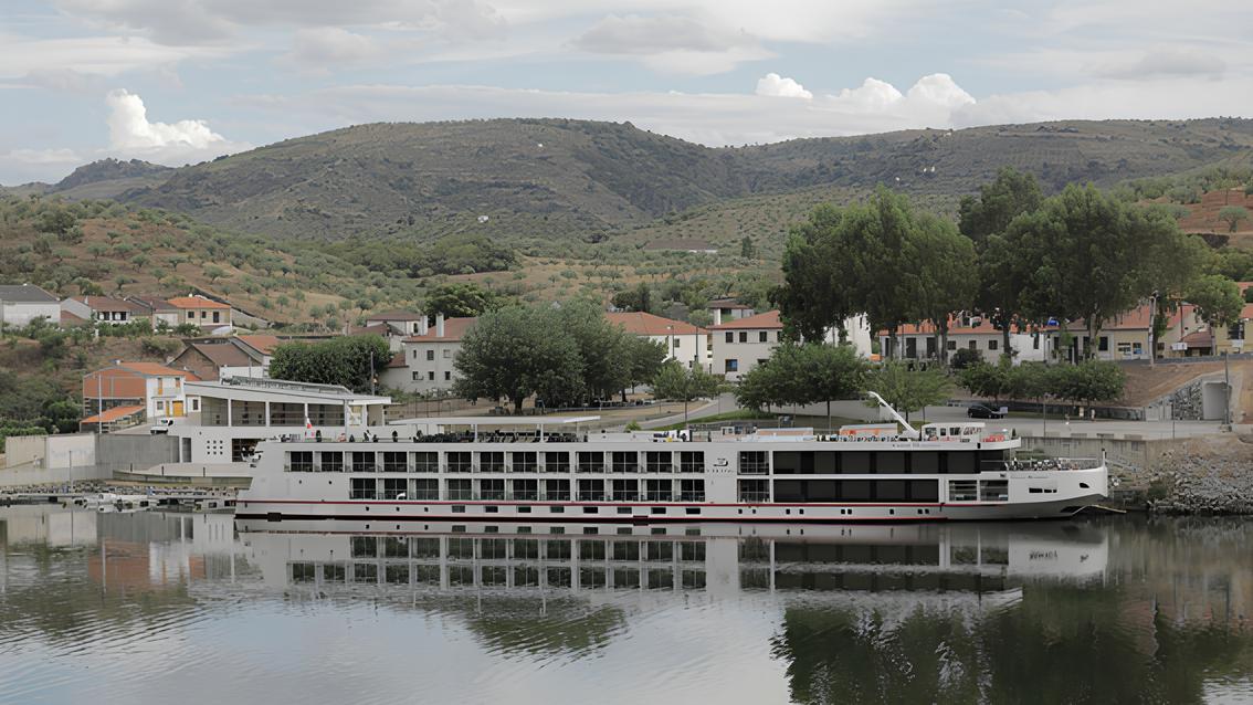Navio de cruzeiro em um rio, com colinas verdes e casas ao fundo em um dia nublado.