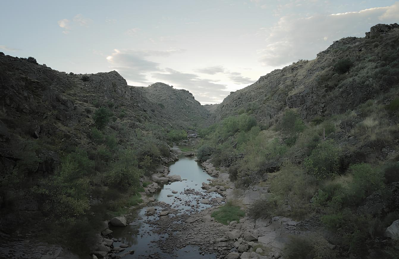 Rio estreito entre montanhas rochosas e vegeta&ccedil;&atilde;o esparsa, sob c&eacute;u nublado.
