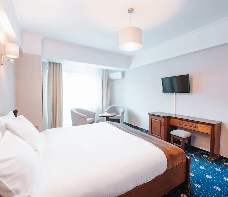 Bright hotel room with a bed, TV, desk, and two chairs near a window.