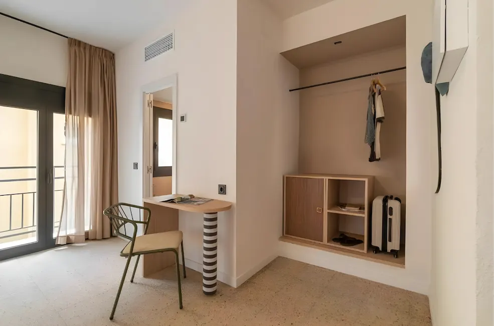 Habitaci&oacute;n minimalista con mesa, silla, armario y maleta junto a una ventana.