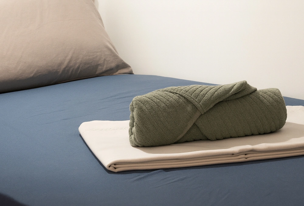 Toalla verde enrollada sobre una cama con s&aacute;banas azules y beige.