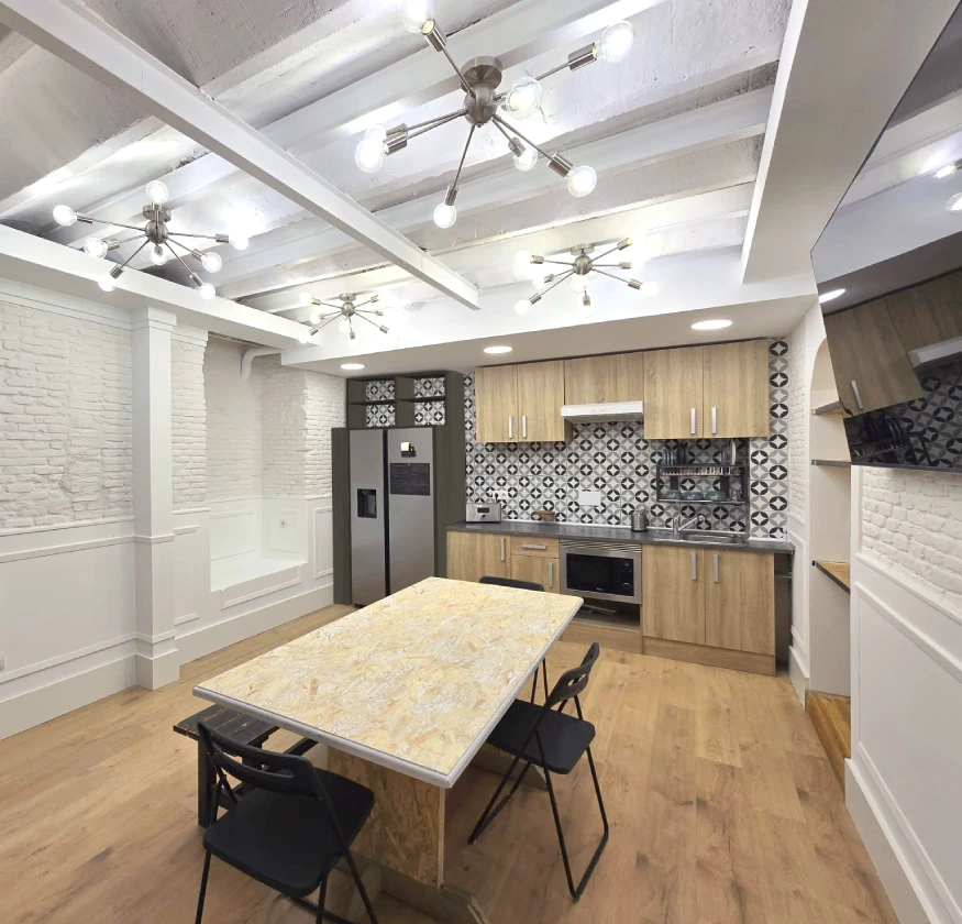 Cocina moderna con mesa central, sillas negras y muebles de madera clara.