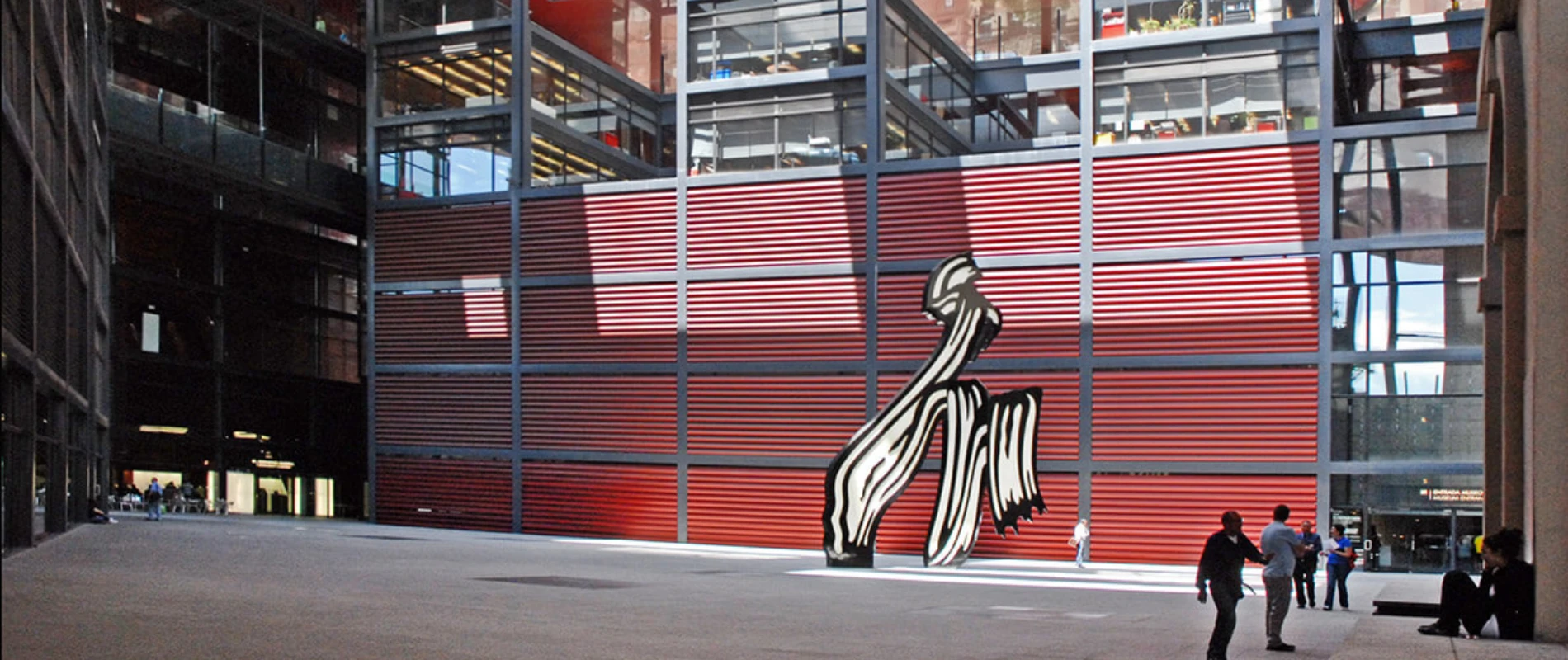 Edificio moderno con fachada roja y escultura blanca; personas en el patio.