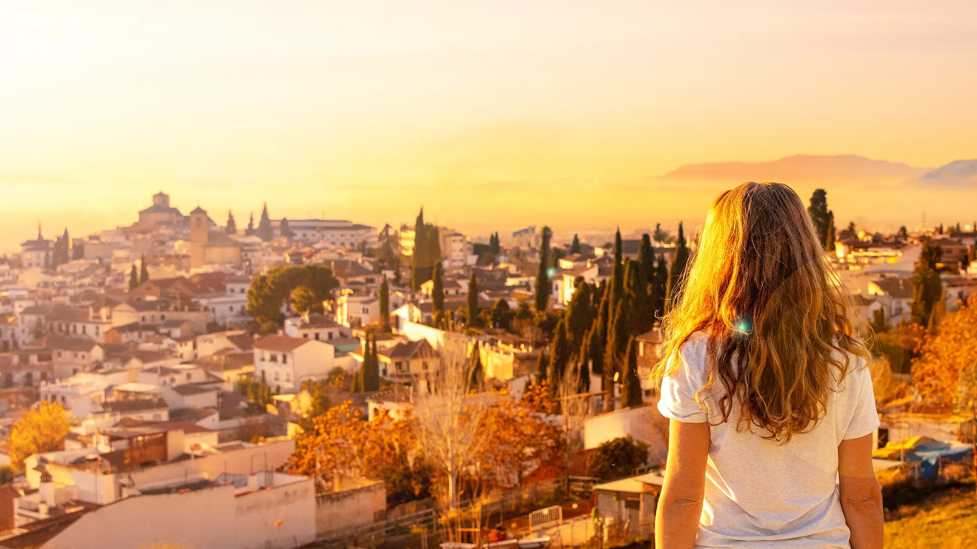 Persona mirando una ciudad al atardecer con &aacute;rboles y edificios.