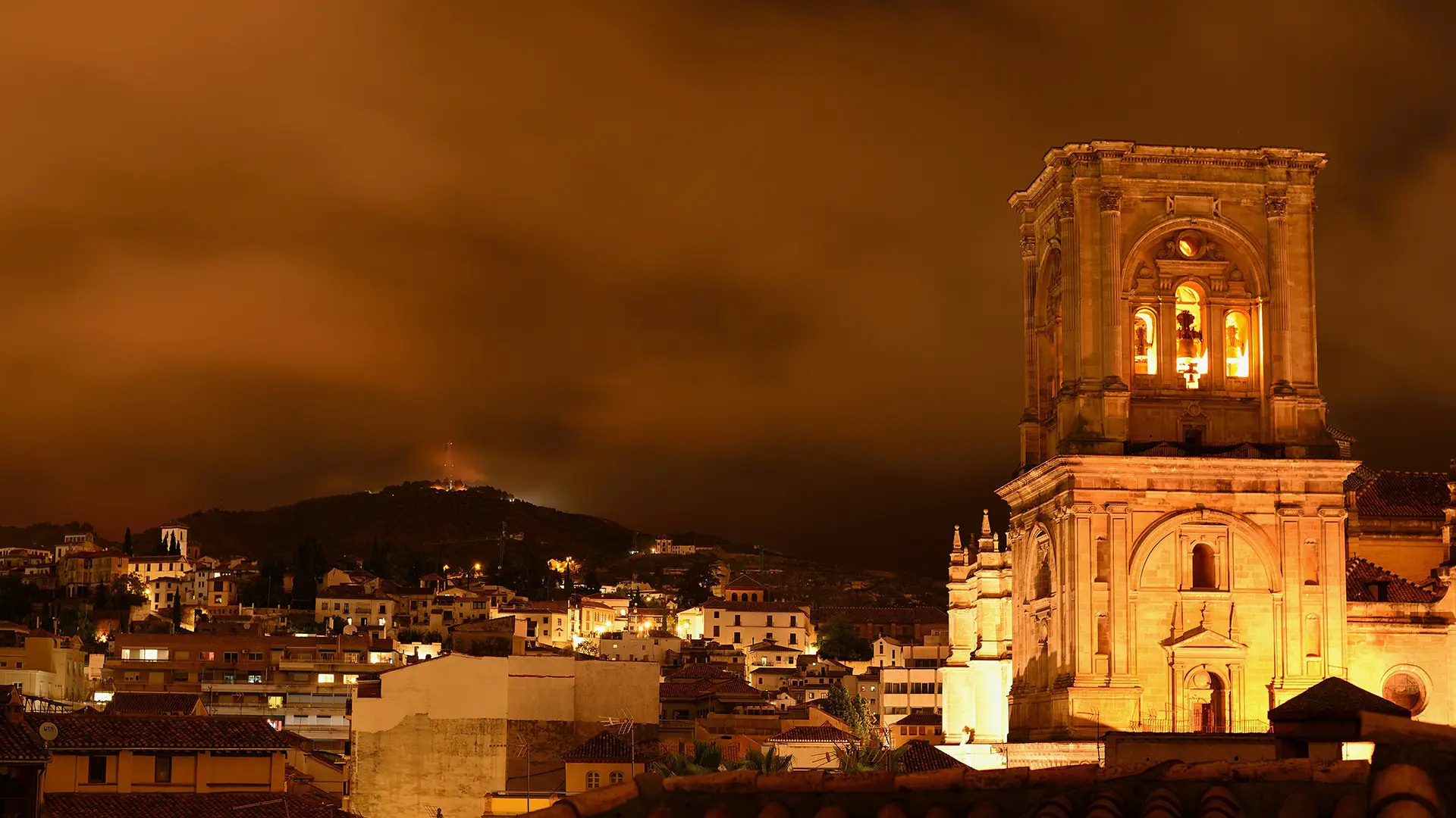 Ciudad iluminada de noche con cielo nublado y torre visible.