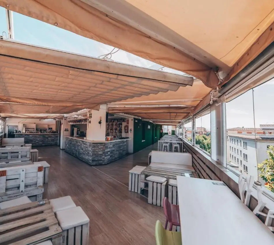 Terraza cubierta con mesas y sillas de palets, vista a la ciudad.