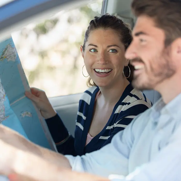 Personas en auto, ella sostiene un mapa y sonr&iacute;e mientras hablan y conducen.