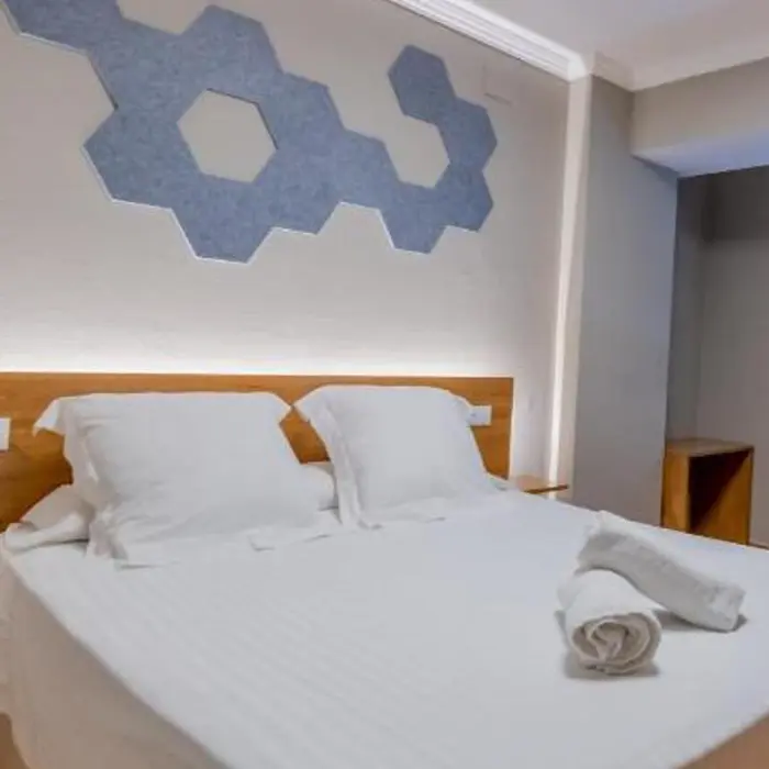 Cama doble con s&aacute;banas y toallas blancas, decoraci&oacute;n de paneles en la pared.