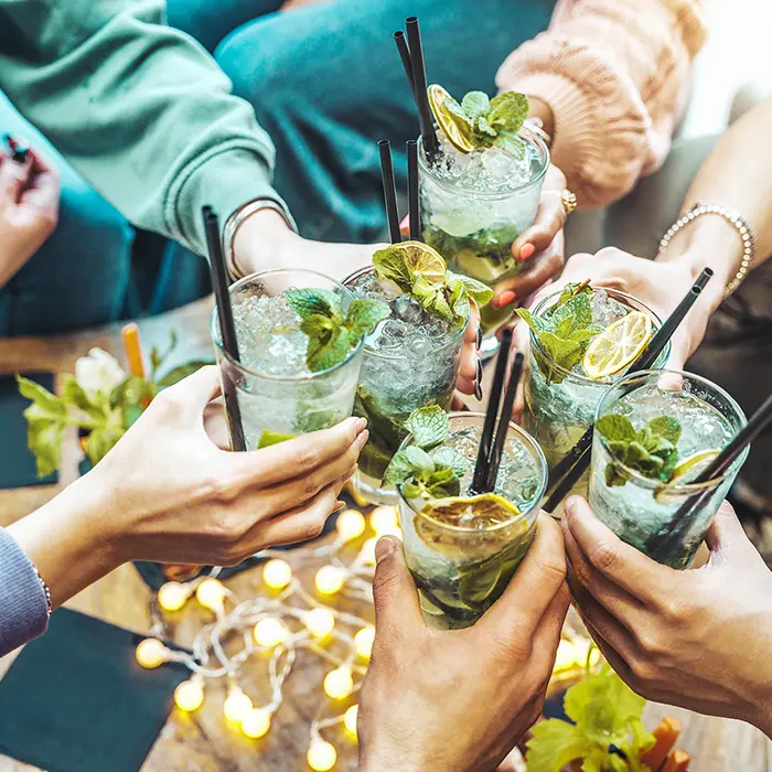 Manos brindando con c&oacute;cteles de menta y lim&oacute;n, luces de fondo.