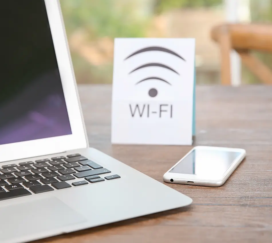 Port&aacute;til y tel&eacute;fono sobre mesa con se&ntilde;al de Wi-Fi al fondo.