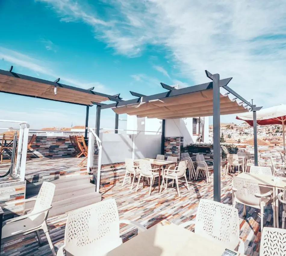 Terraza con mesas y sillas bajo p&eacute;rgolas, cielo azul despejado.