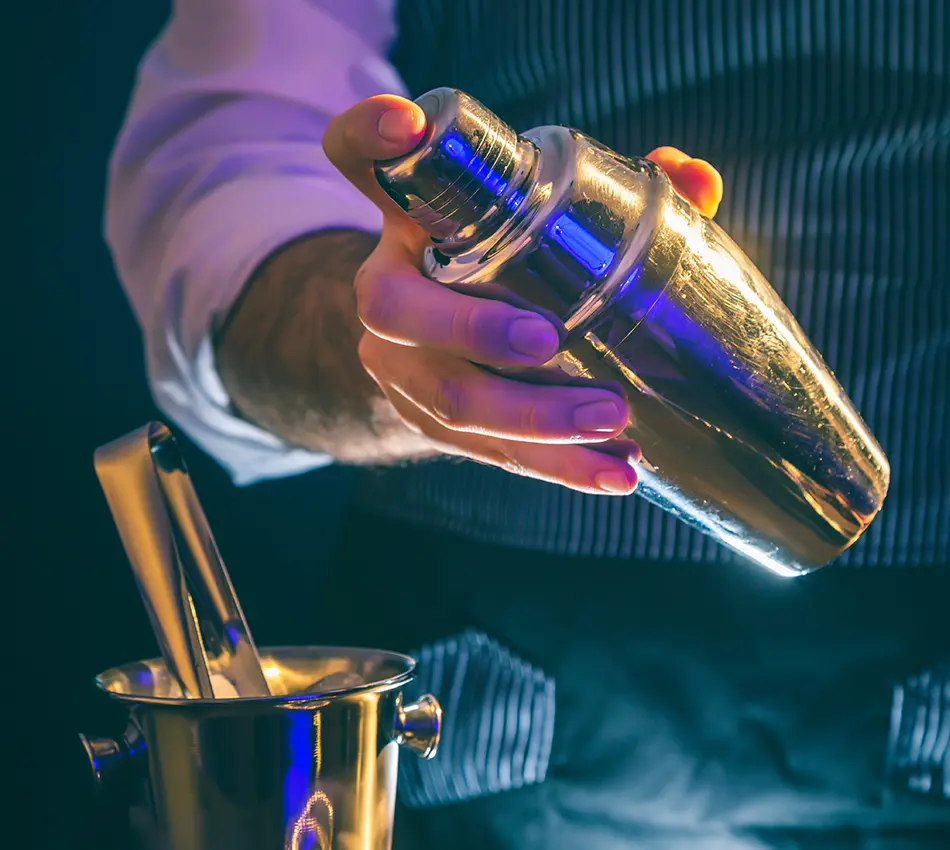 Mano sosteniendo una coctelera met&aacute;lica brillante junto a un cubo con pinzas.