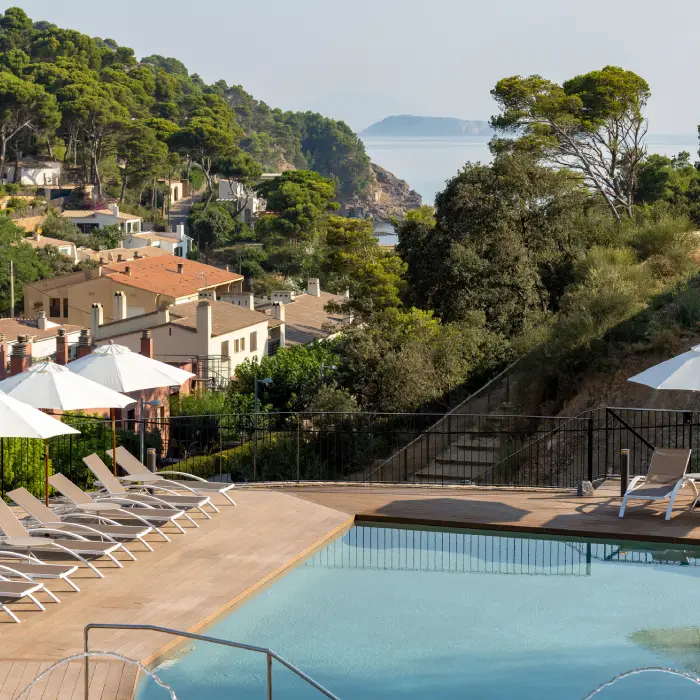 Piscina con tumbonas y sombrillas, fondo de casas y paisaje boscoso junto al mar.