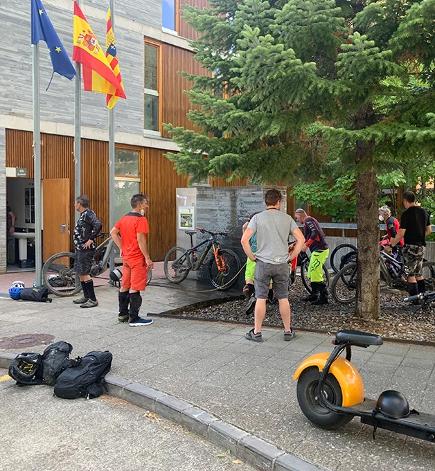 Ciclistas conversan frente a un edificio con banderas y bicicletas estacionadas.