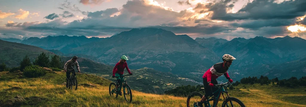 Ciclistas montando en monta&ntilde;a al atardecer con nubes y monta&ntilde;as al fondo.