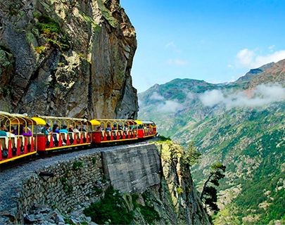 Tren rojo y amarillo recorre una monta&ntilde;a con paisaje verde y cielo azul.