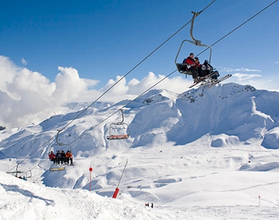 Esquiadores en telesilla sobre monta&ntilde;as nevadas bajo un cielo azul.