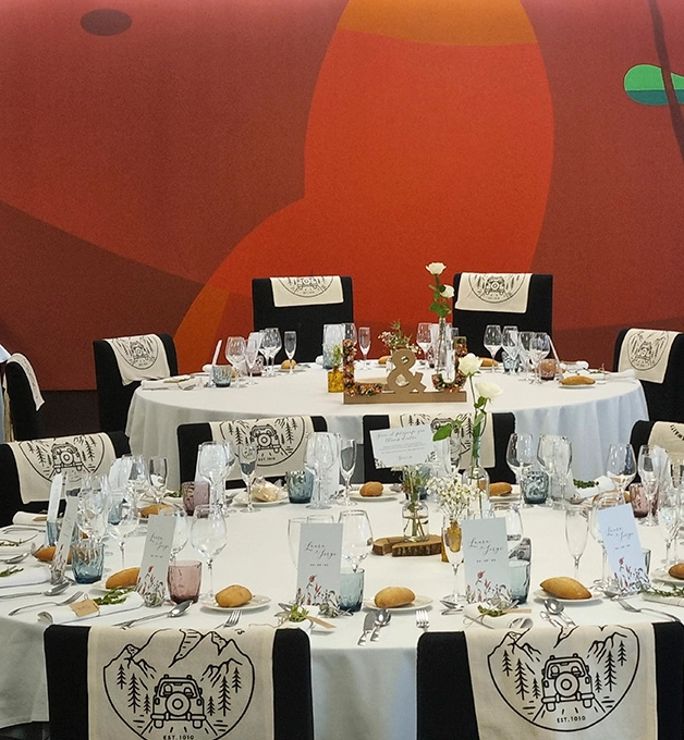 Mesa redonda decorada para evento, con copas y flores. Fondo colorido.