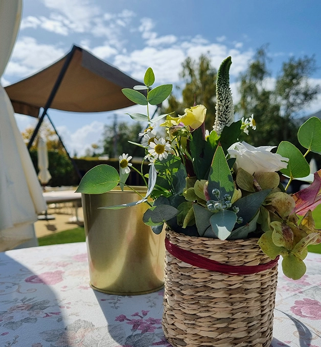 Ramo de flores en maceta tejida, mesa de jard&iacute;n, cielo azul y sombrilla.