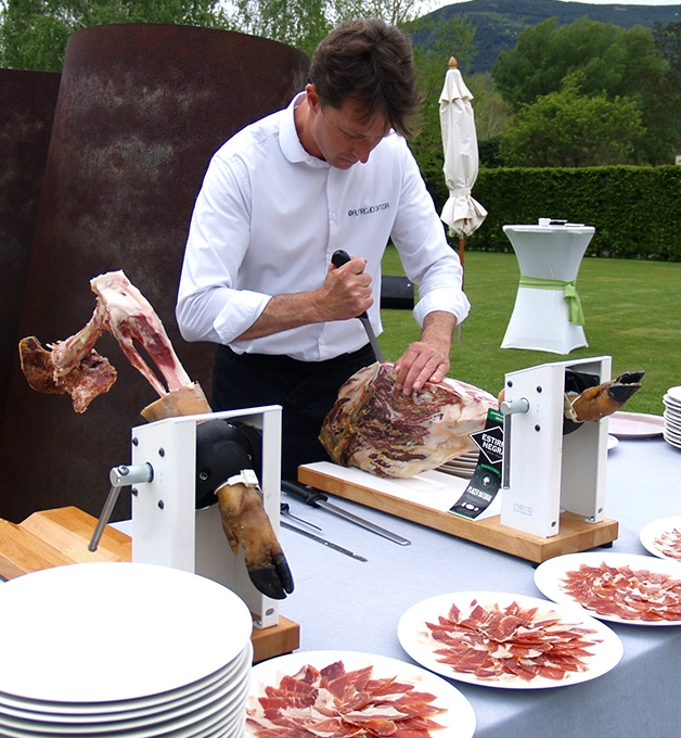 Persona cortando jam&oacute;n en una mesa con platos de lonchas al aire libre.