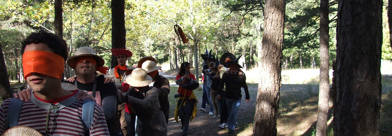 Personas con antifaces naranjas caminan en fila en el bosque, gui&aacute;ndose mutuamente.