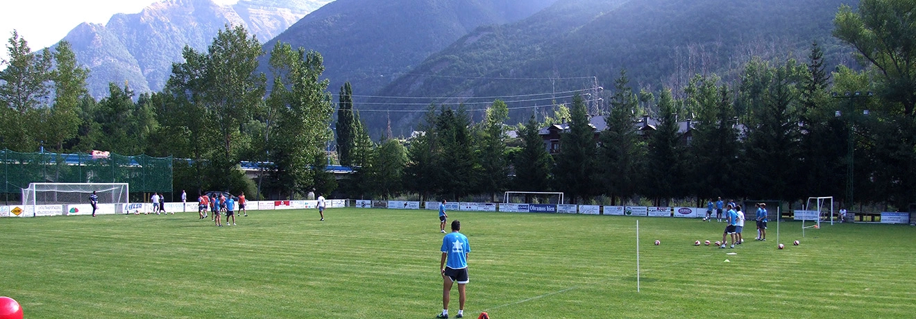 Jugadores entrenan en una cancha de f&uacute;tbol rodeada de monta&ntilde;as y &aacute;rboles.