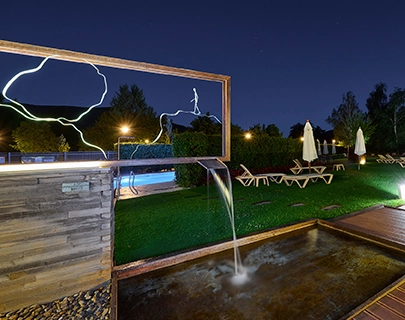 Paisaje nocturno con fuente y sillones junto a piscina iluminada.