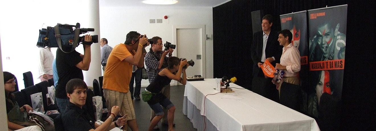 Rueda de prensa con fot&oacute;grafos y c&aacute;maras frente a dos personas con carteles.