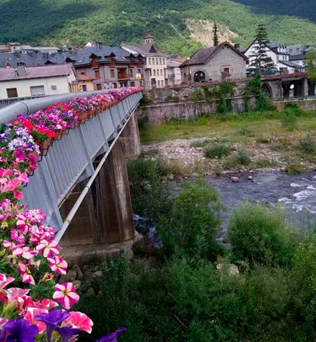 Puente con flores coloridas y r&iacute;o, rodeado de casas y monta&ntilde;as verdes.