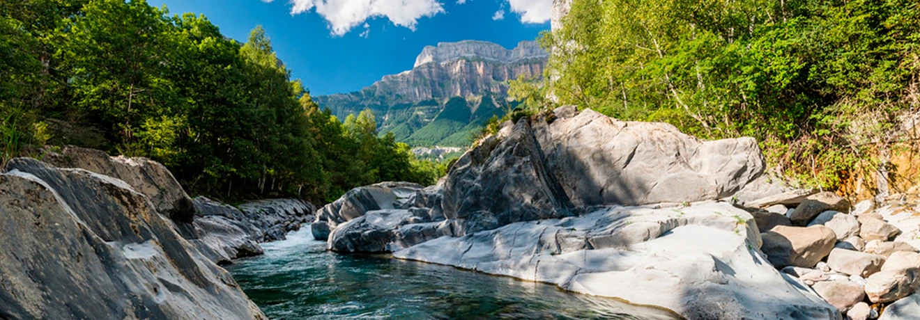 R&iacute;o rodeado de &aacute;rboles y monta&ntilde;as bajo un cielo azul con nubes.