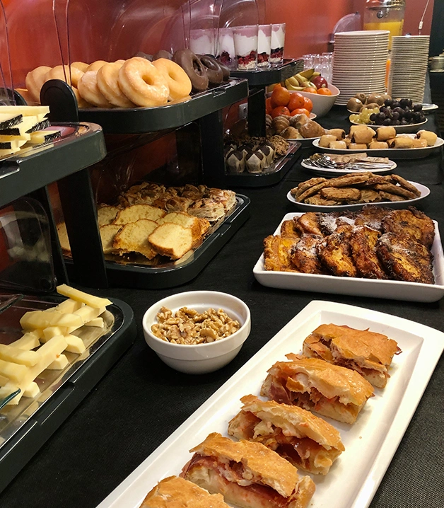 Buf&eacute; de desayuno con panes, frutas y postres variados sobre una mesa.