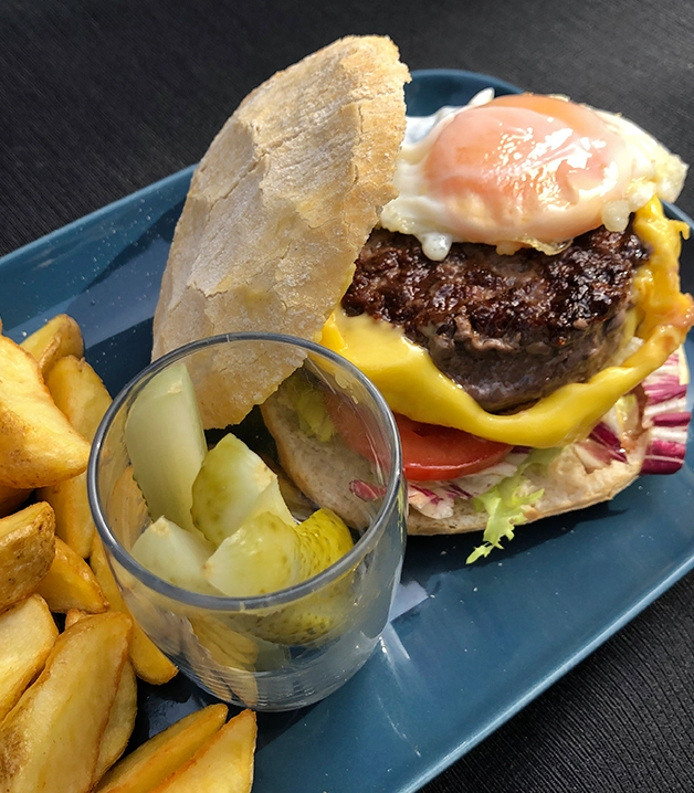 Hamburguesa con queso y huevo, papas fritas y pepinillos en plato azul.