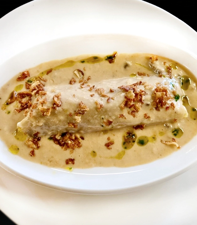 Tamale ba&ntilde;ado en salsa cremosa con cebolla crujiente en un plato blanco.