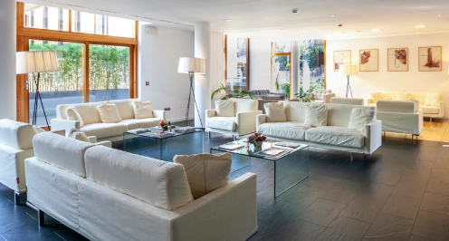 Sala moderna con sof&aacute;s blancos, mesas de cristal y grandes ventanas.