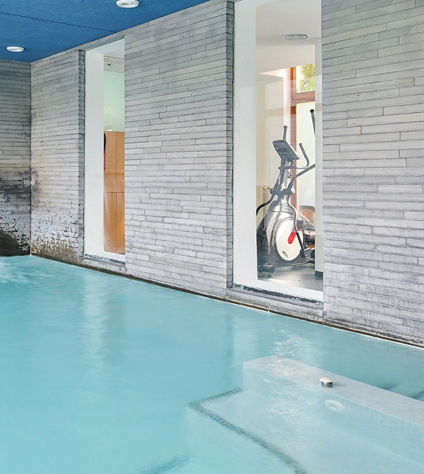 Piscina interior junto a una sala con bicicleta est&aacute;tica detr&aacute;s de una pared de ladrillo.