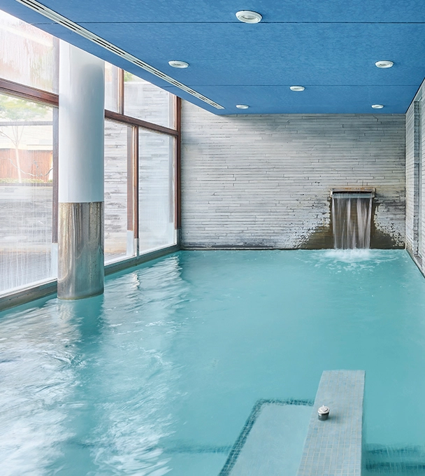 Piscina interior con cascada, ventanas grandes y techo azul.