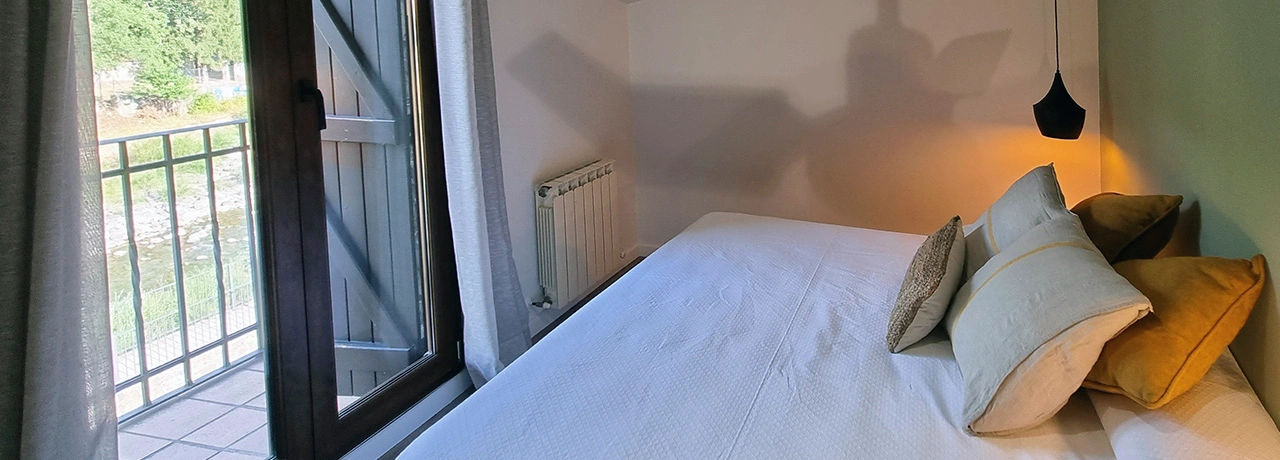 Habitaci&oacute;n con cama, ventana y luz suave sobre cojines coloridos.