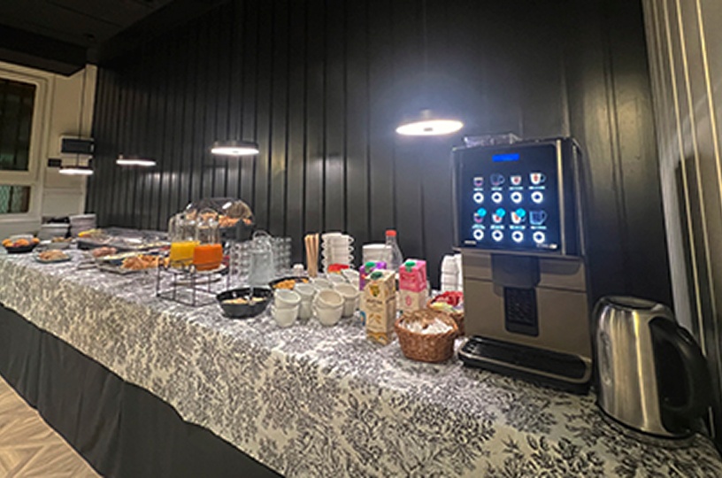 Mesa de buffet con m&aacute;quina de caf&eacute; y vajilla bajo luz blanca.
