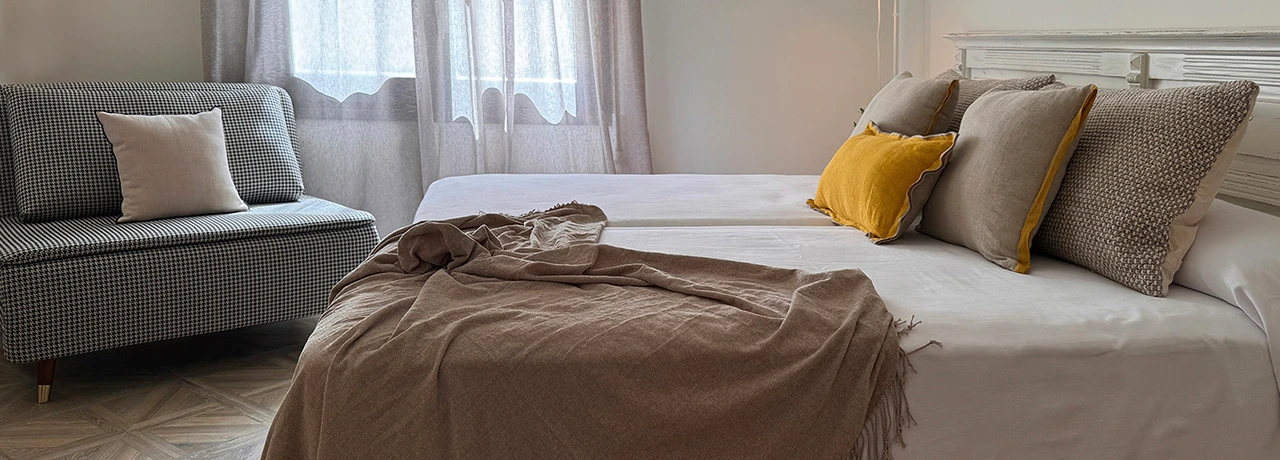 Cama con cojines y manta, junto a un sofá, en una habitación luminosa.