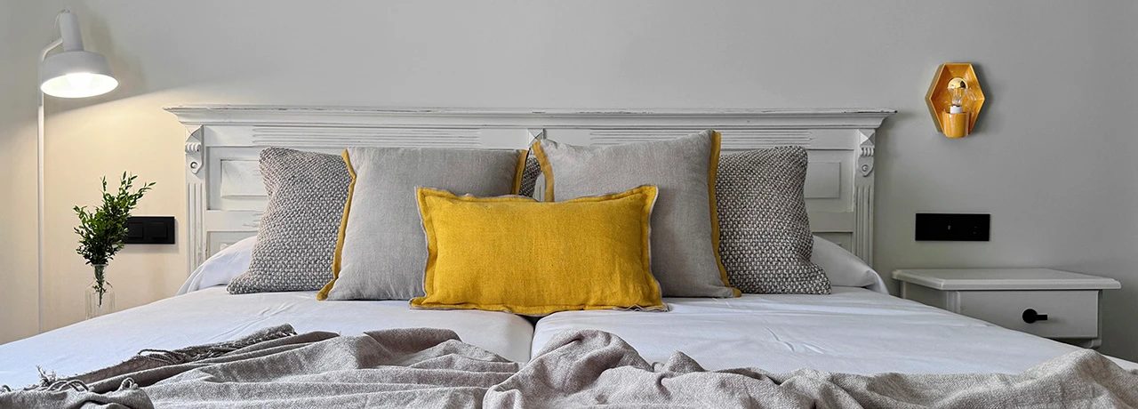 Cama con cojines grises y amarillos, lámpara de pared y mesa de noche blanca.